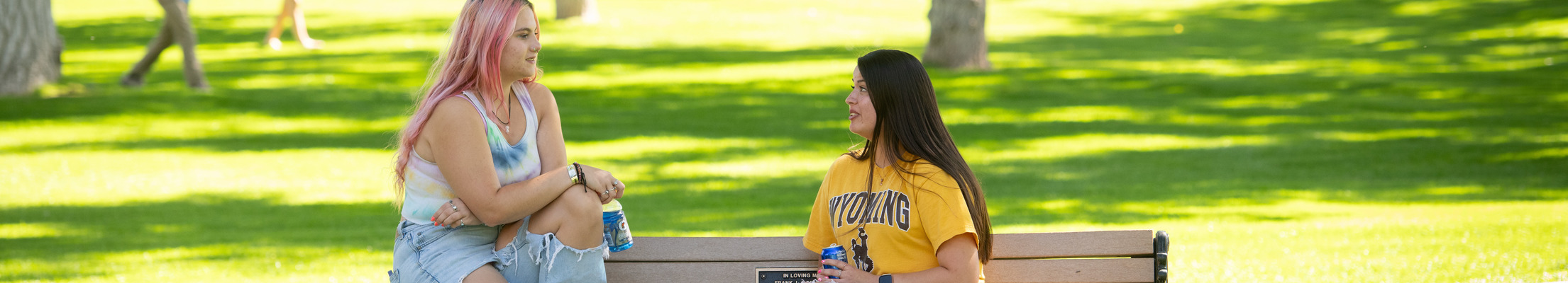 Two students sit on a bench on campus talking.