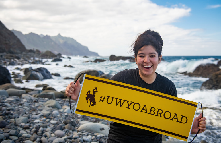a student poses in front of the ocean with a UW study abroad banner. 