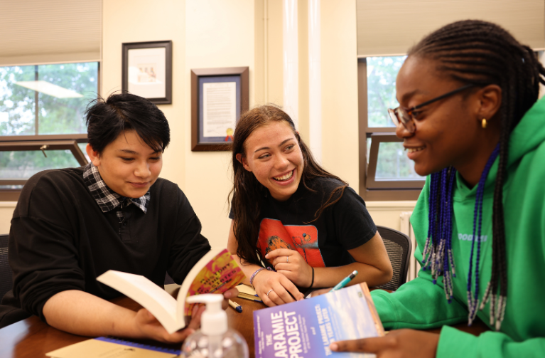 A diverse group of smiling students sitting at a table while holding books, laughing, and interacting with each other