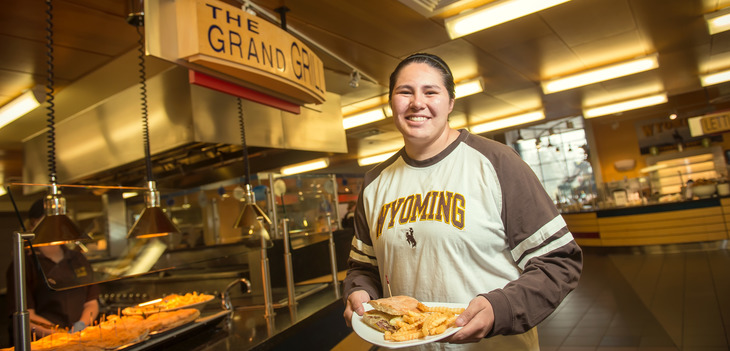 Person smiling and holding plate of food in front of The Grand Grill at Washakie Dining Center